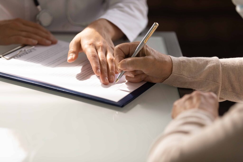 Person signing legal document with advisor’s guidance, representing consent form or estate planning agreement. Person signing legal document with advisor’s guidance, representing consent form or estate planning agreement.